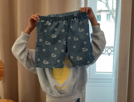 Atelier couture enfant : une enfant pose fièrement, cachant son visage derrière un short en jean à motifs de fleurs blanches qu'elle a cousu elle-même.