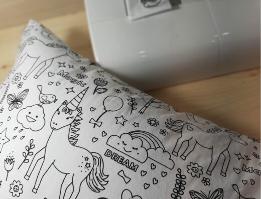 Atelier couture : un coussin à colorier avec motifs de licornes, nuages et papillons, posé devant une machine à coudre, pour un loisir créatif enfant.