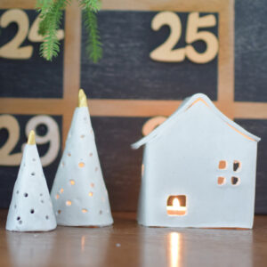 Maison et sapins de Noël en céramique blanche avec bougie allumée devant un calendrier de l'avent mural en bois.