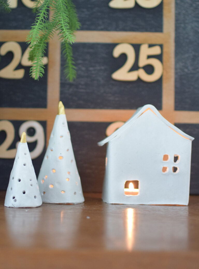 Maison et sapins de Noël en céramique blanche avec bougie allumée devant un calendrier de l'avent mural en bois.