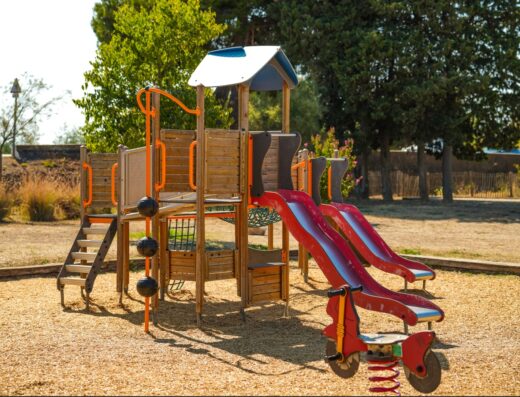 Aire de jeux extérieure en bois pour enfants avec toboggan rouge double, échelle et jeu sur ressort sur sol amortissant en copeaux.