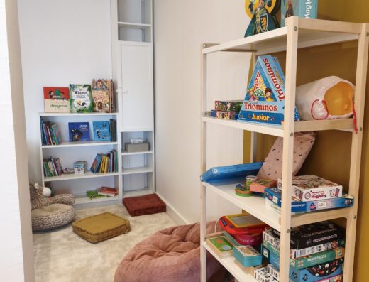 Coin lecture enfant avec étagères de livres, jeux de société, tapis doux et pouf rose dans une salle de jeux lumineuse.