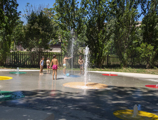 Enfants jouant dans un parc de jeux d'eau avec jets verticaux et dalles colorées en plein air.