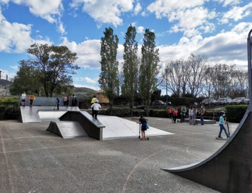 Enfants sur des trottinettes dans un skatepark extérieur avec rampes métalliques et arbres en arrière-plan sous un ciel nuageux.