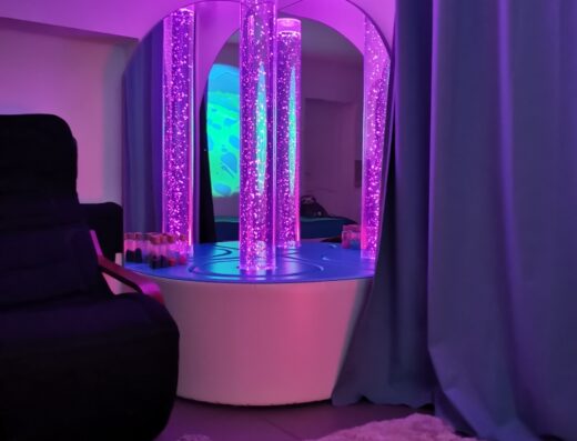 Colonne à bulles lumineuse violette dans une salle multisensorielle Snoezelen avec tapis doux et fauteuil de relaxation.