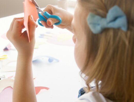 Petite fille découpant du papier orange avec des ciseaux bleus lors d'un atelier loisirs créatifs.