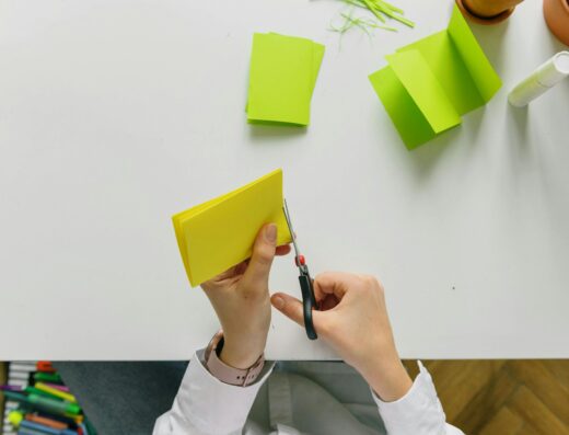 Une personne découpe du papier jaune plié avec des ciseaux sur un bureau blanc lors d'un atelier loisirs créatifs