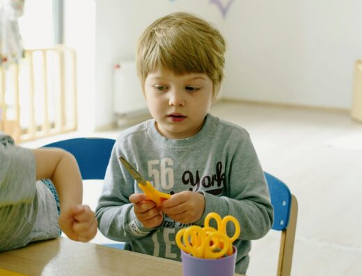 Enfant assis à une table de classe découpant du papier avec des ciseaux jaunes lors d'une activité manuelle en école maternelle.