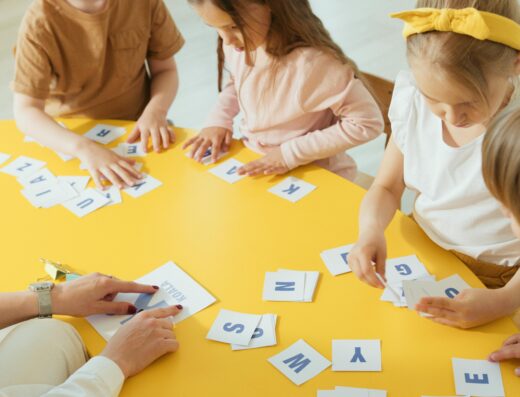 Des enfants apprennent les lettres de l'alphabet autour d'une table jaune avec une institutrice